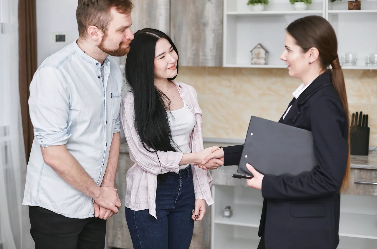 A real estate agent shakes hands with a smiling young couple in their kitchen